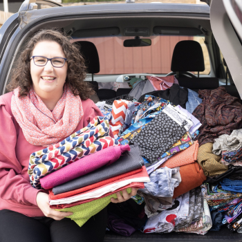 Smiling woman holds a large stack of colourful fabric in front of a car filled with more textiles, ready for reuse or crafting.